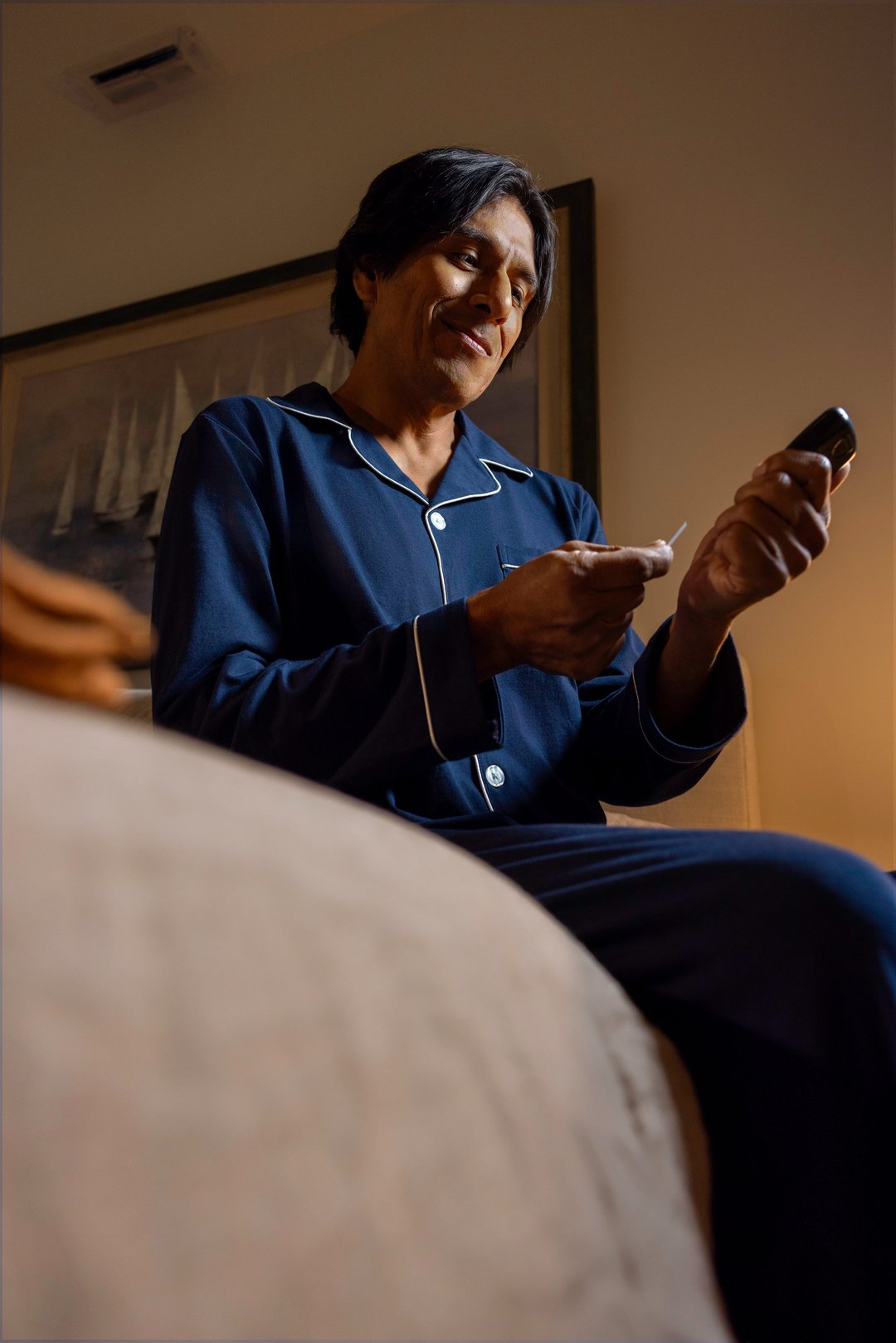 Man in navy pajamas sitting on the edge of his bed at night, smiling as he checks his blood sugar with a Contour meter and test strip